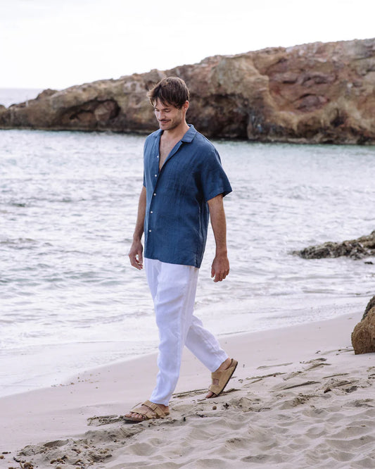 Man walking on a beach wearing a blue shirt and white pants.