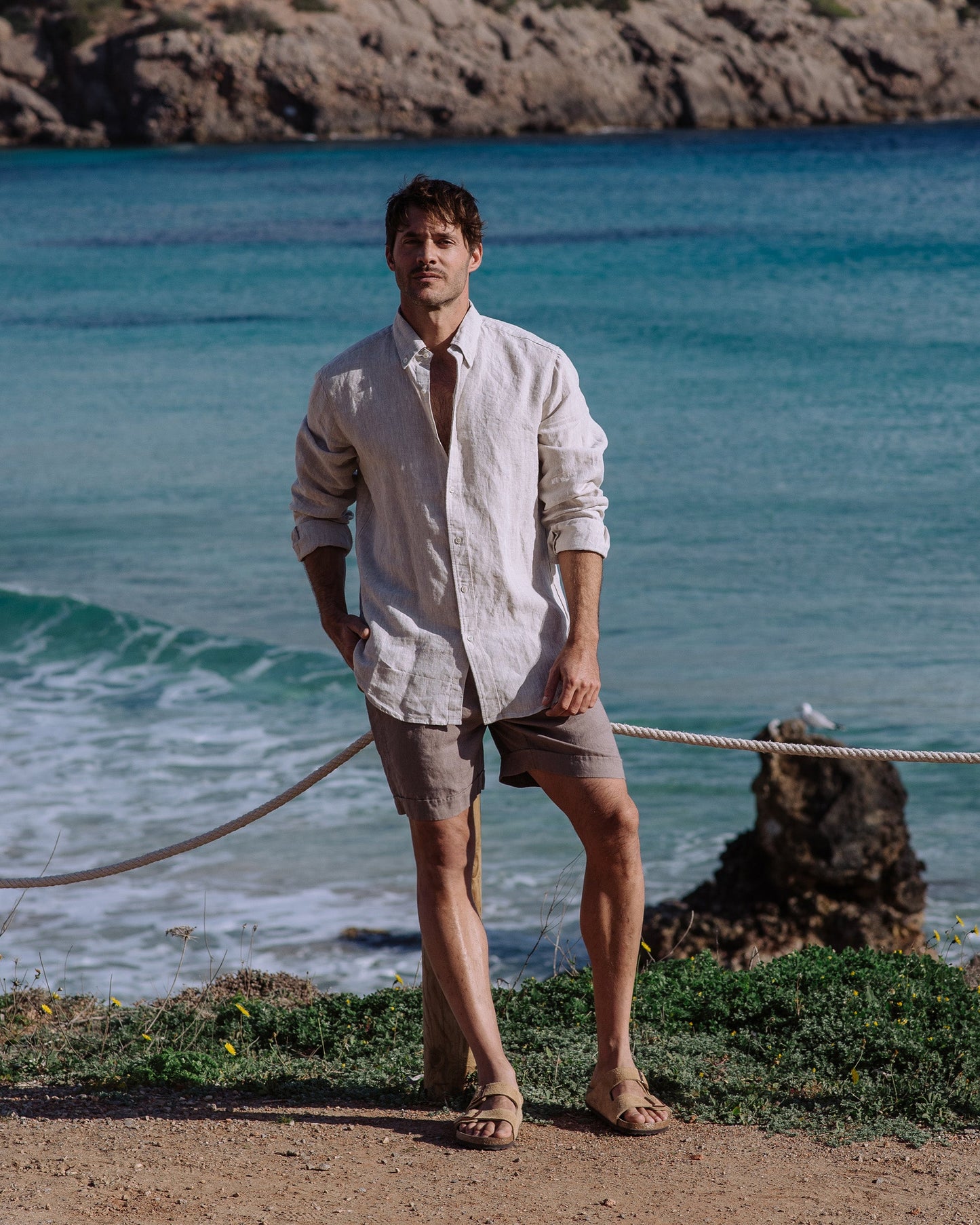 Man standing by the ocean wearing a light shirt and shorts.