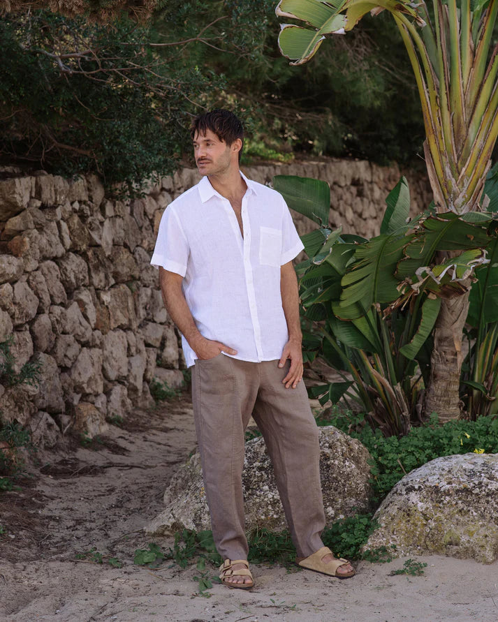 Man in a white shirt and beige pants standing in a natural setting with stone wall and plants.