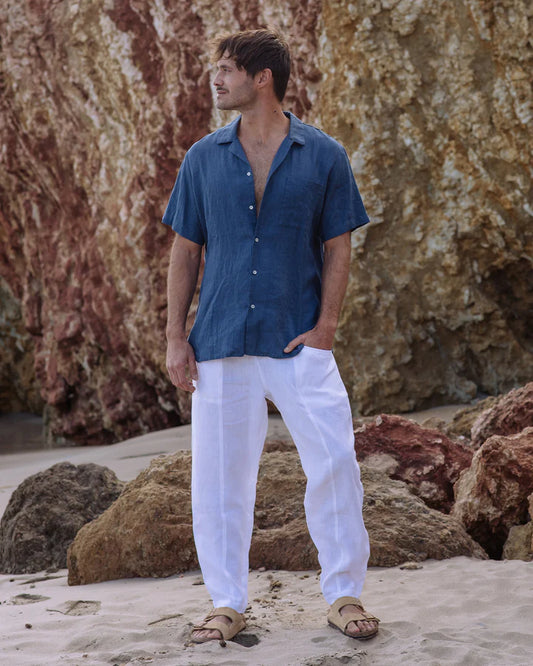 Man wearing a blue shirt and white pants standing on a rocky beach.