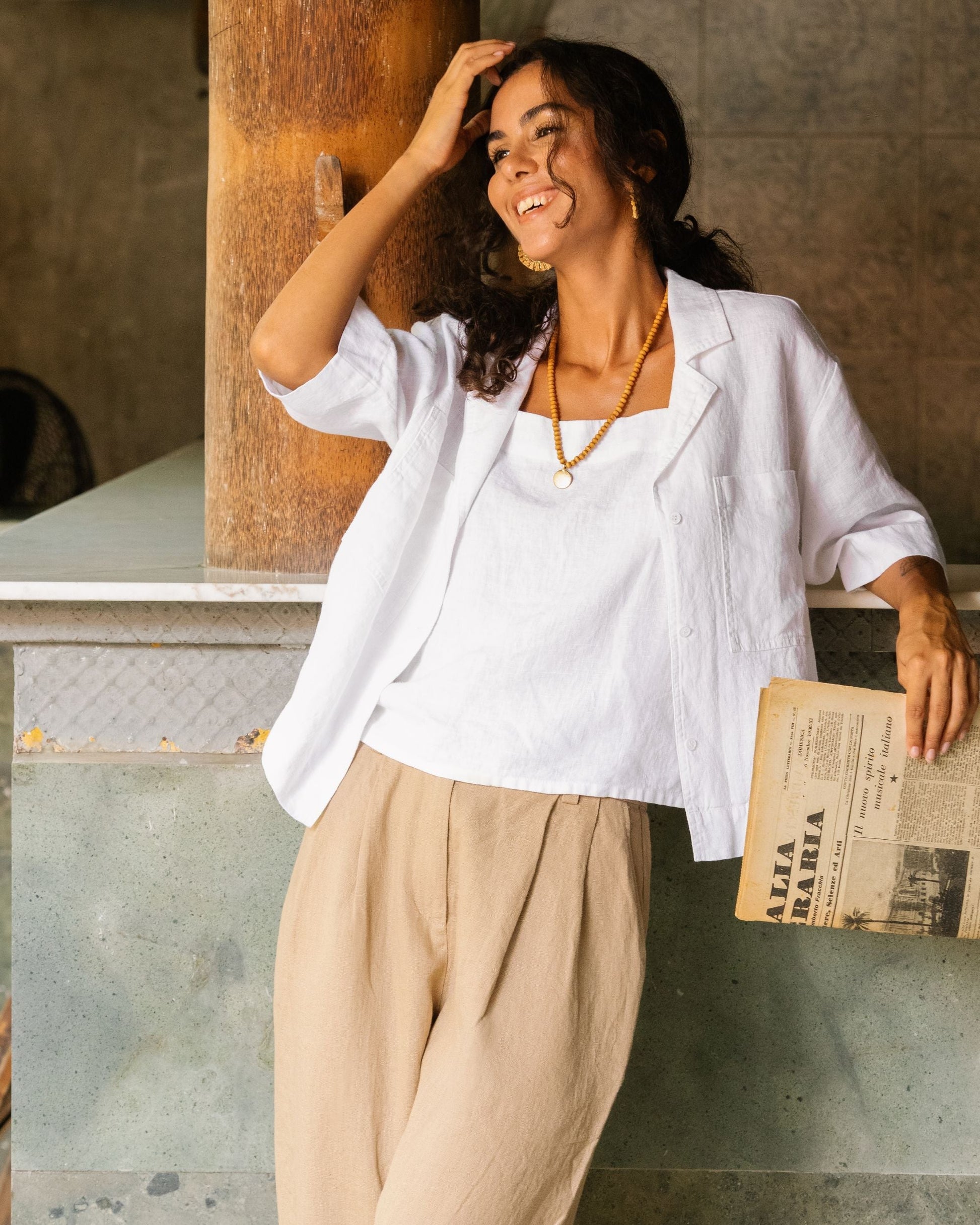 Woman in a white blazer and beige pants holding a newspaper, standing against a rustic wall.