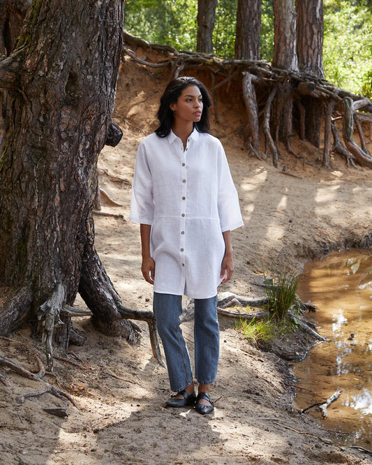 Person wearing a white shirt and blue jeans standing in a natural setting with trees and water.